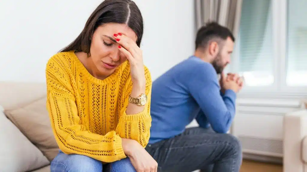 A woman in a yellow sweater holds her forehead while a man sits blurred behind her.