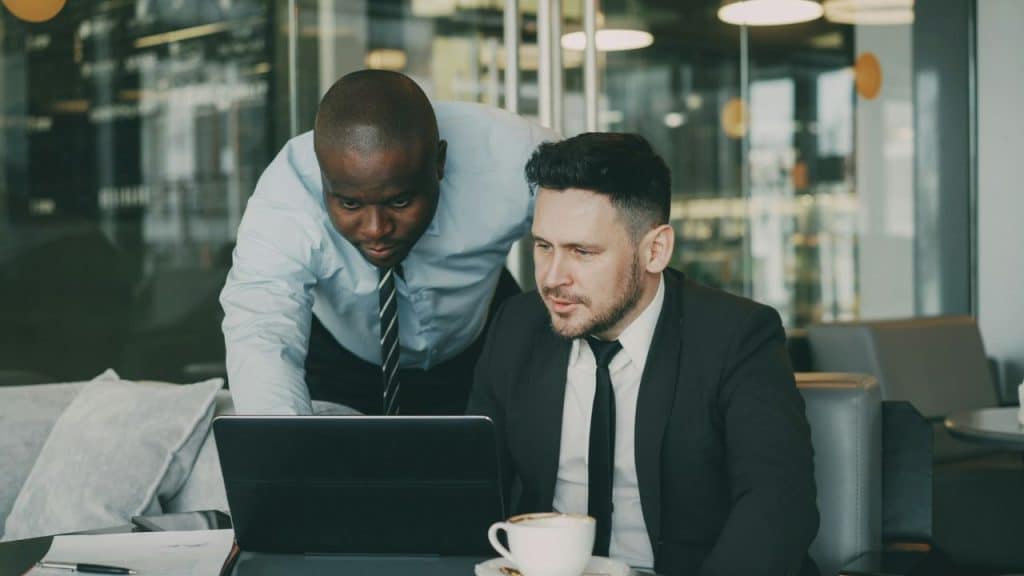Two men in business attire look at a laptop screen together in an office setting.