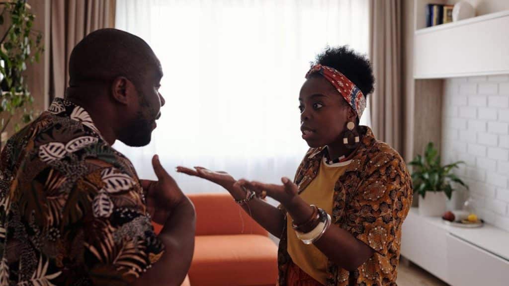 A man and a woman, both wearing patterned shirts, stand indoors facing each other and are gesturing during an apparently heated argument.