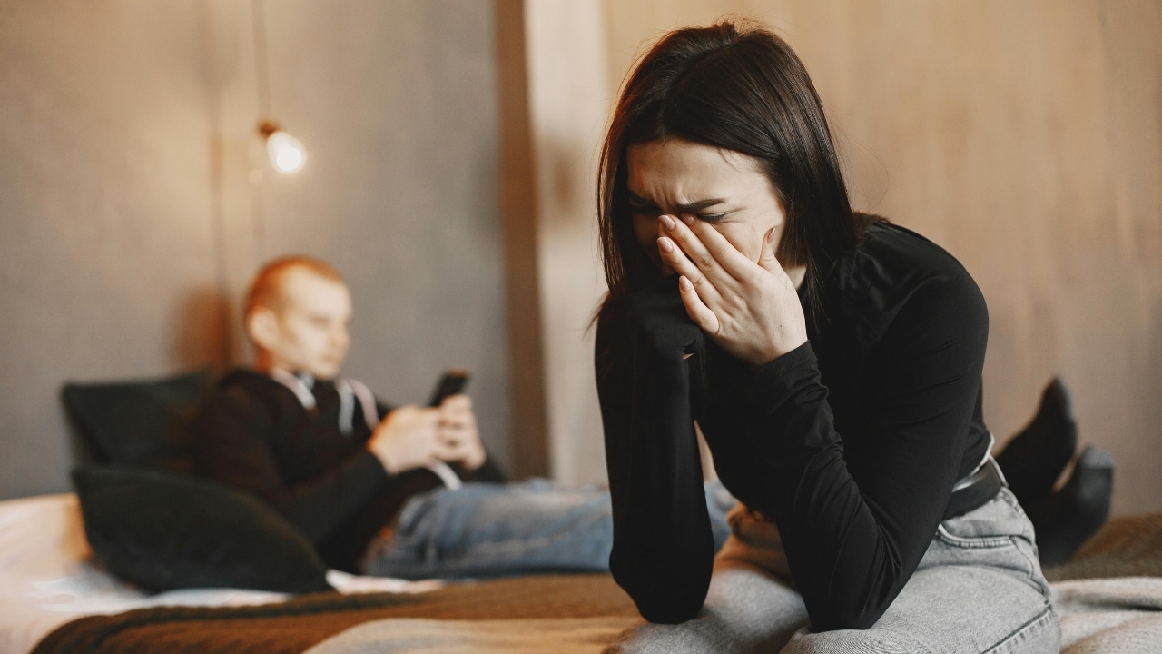 A woman sits on a bed crying with her hands over her face while a man uses a phone.