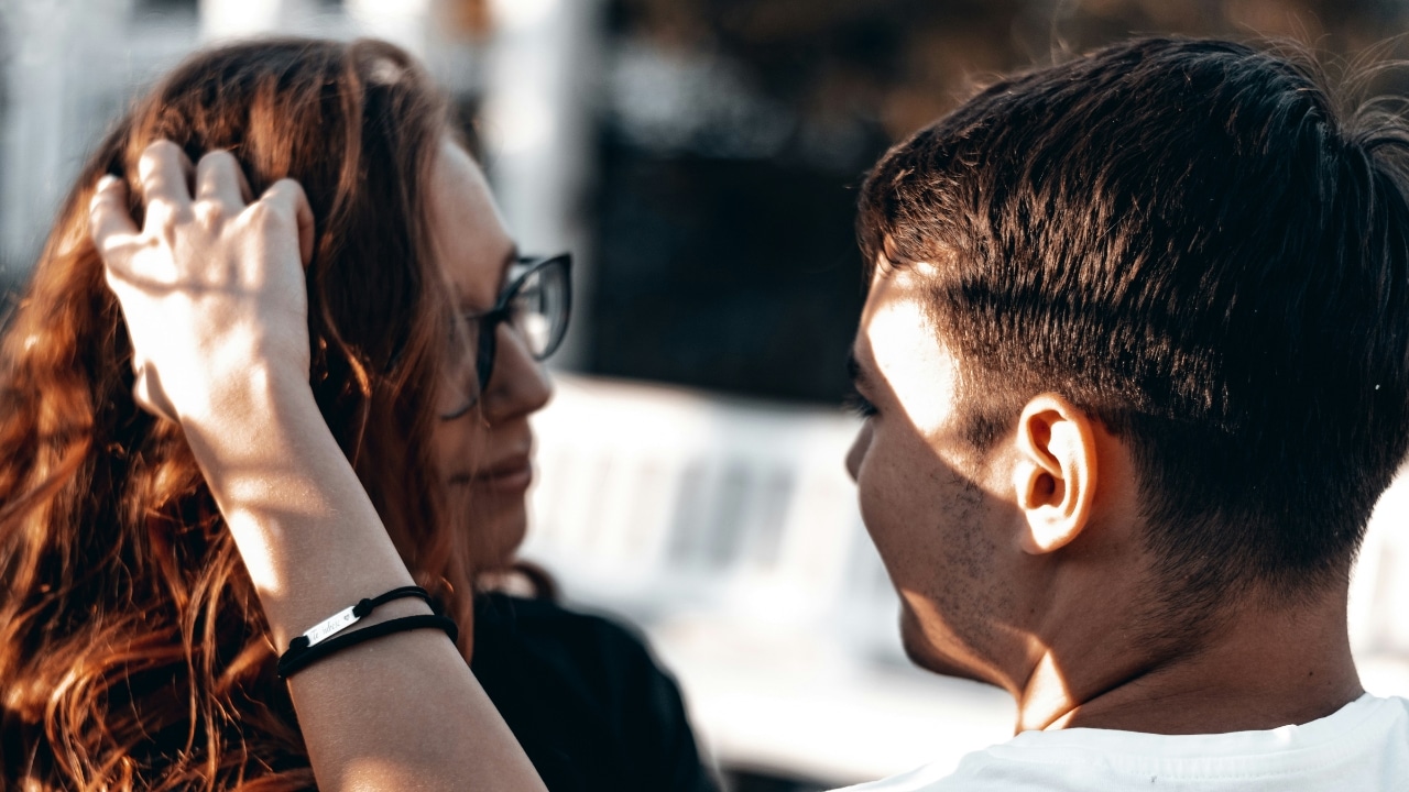 A person's hand touches a woman’s hair while she looks at a man outdoors.