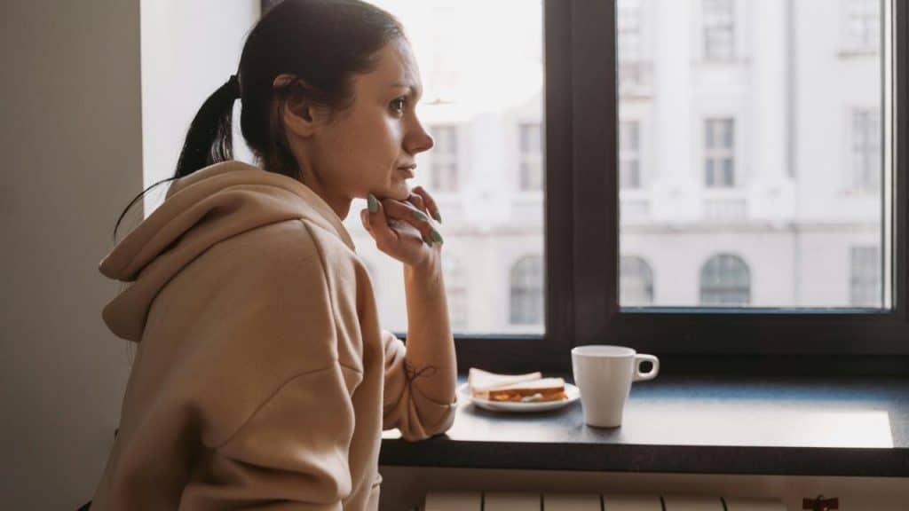 A woman in a hoodie gazes out a window beside a cup of coffee and a small plate.