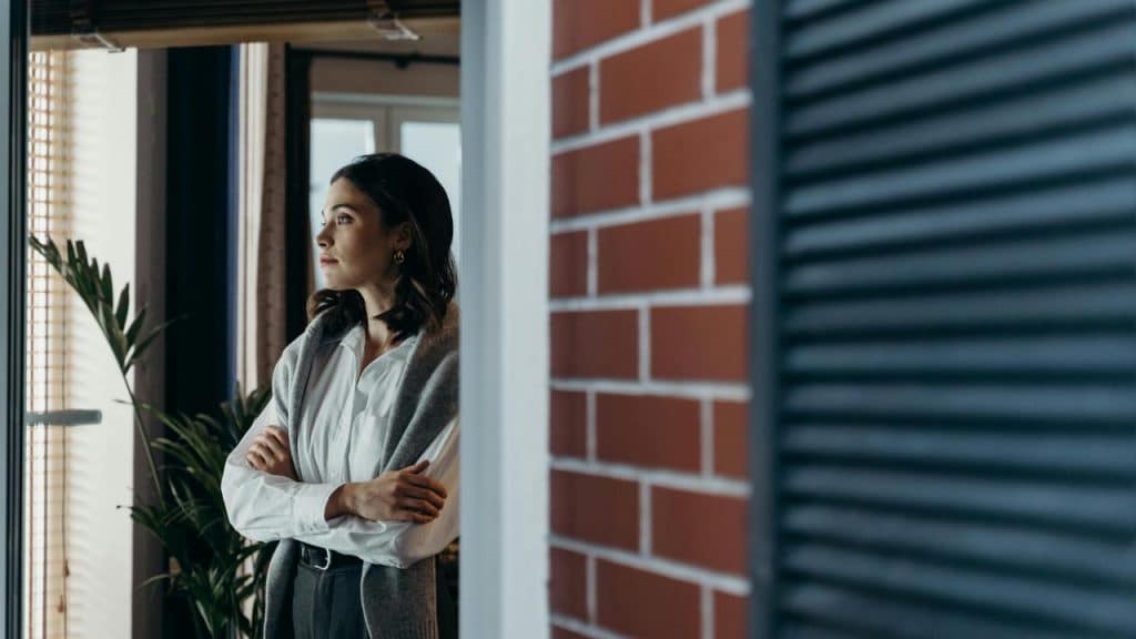 A person standing by a window with folded arms, looking outside.