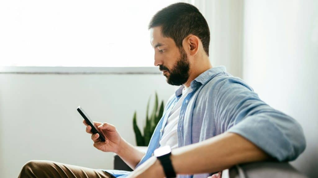 A bearded man in a light blue shirt sits indoors and looks at his smartphone.