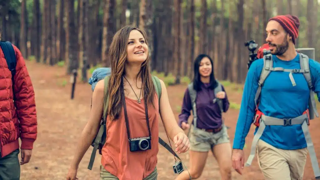 Four people with backpacks walk through a sunlit forest trail while one woman looks upward.