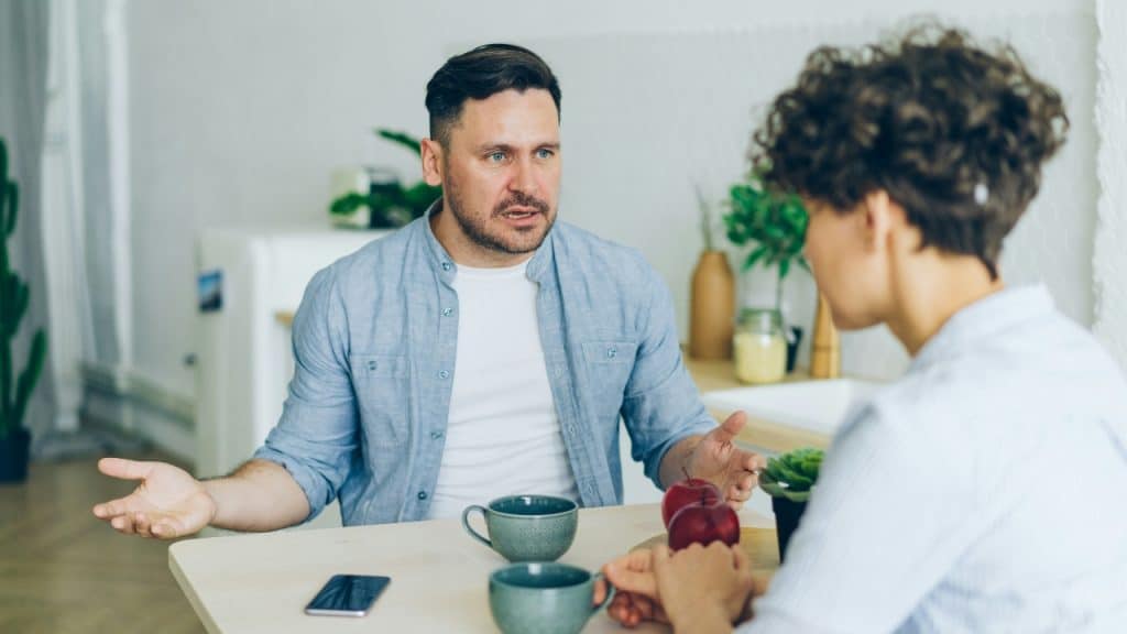 A man sitting at a table gesturing with open hands while speaking to a curly-haired woman.
