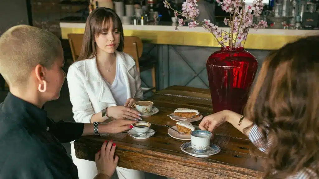 Three women sit at a wooden table with coffee, cake, and a red vase.