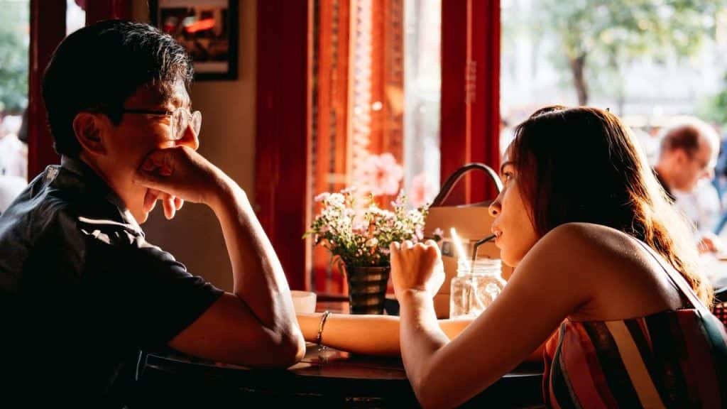 A man and woman sit at a sunlit cafe table, looking directly at each other.