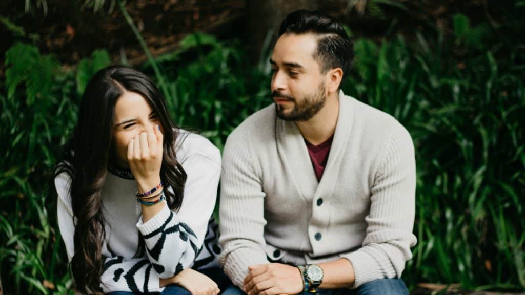 A woman covers her mouth while laughing next to a man sitting in front of greenery.