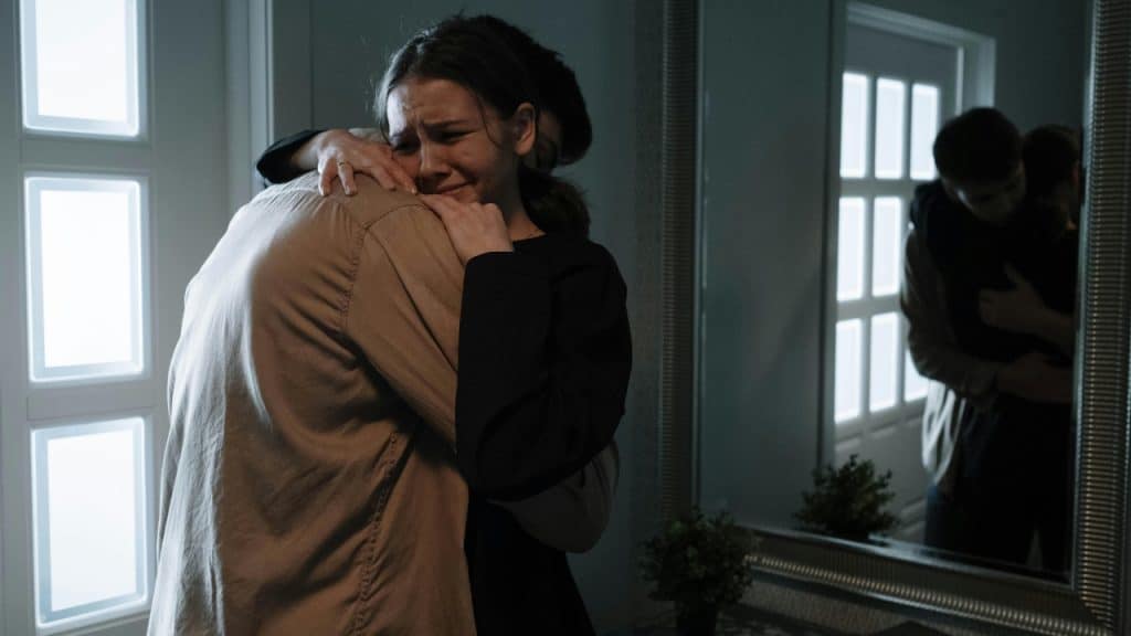 A woman cries while hugging a man in a dimly lit room near a mirror.