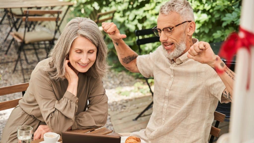 An older man with tattoos and glasses gestures while sitting with a woman at a table.