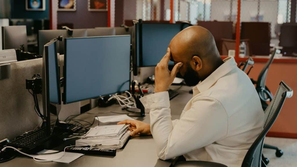 A man sits at an office desk with his hand on his forehead looking at monitors.