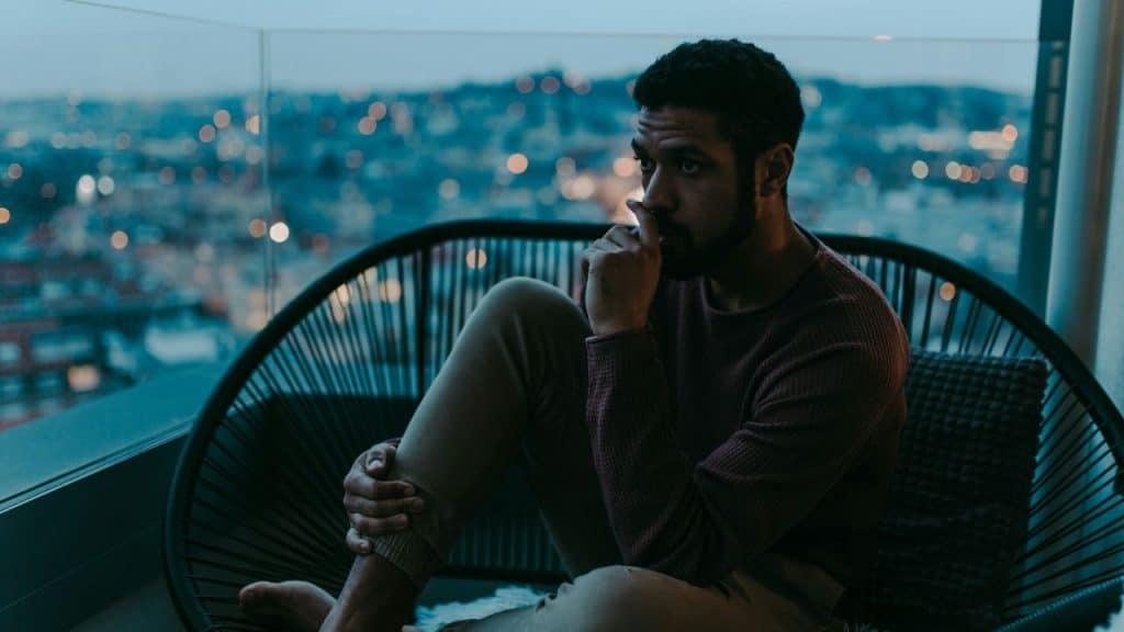 A man sits on a balcony chair at dusk, looking out at city lights.
