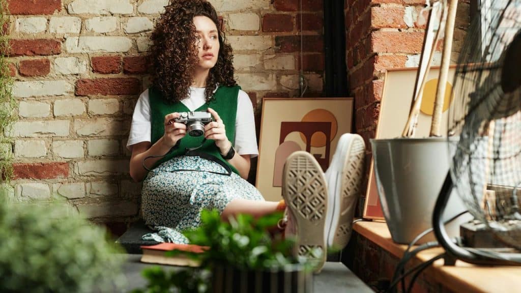 A woman sitting against a brick wall holding a camera in a cozy room.