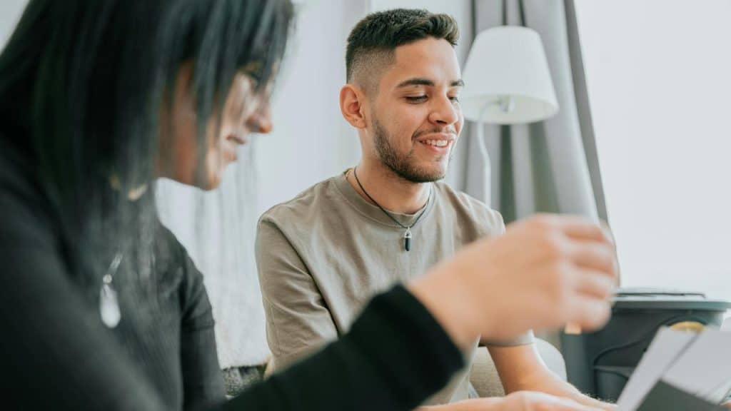 A smiling man sitting with another person, looking at something together indoors.