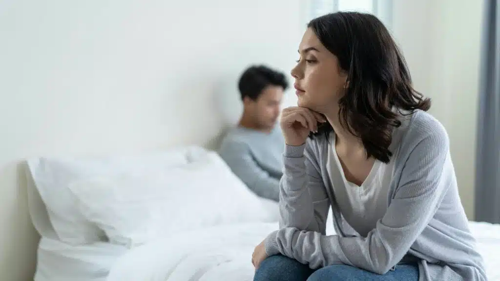 A woman sits on a bed looking away while a blurred man sits in the background.