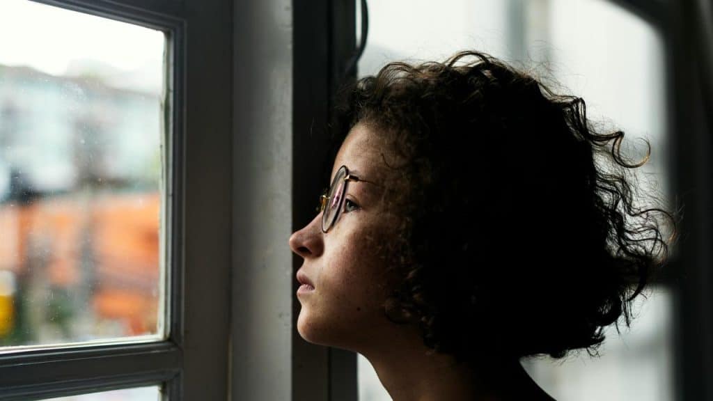 A person with glasses looking out a window thoughtfully.