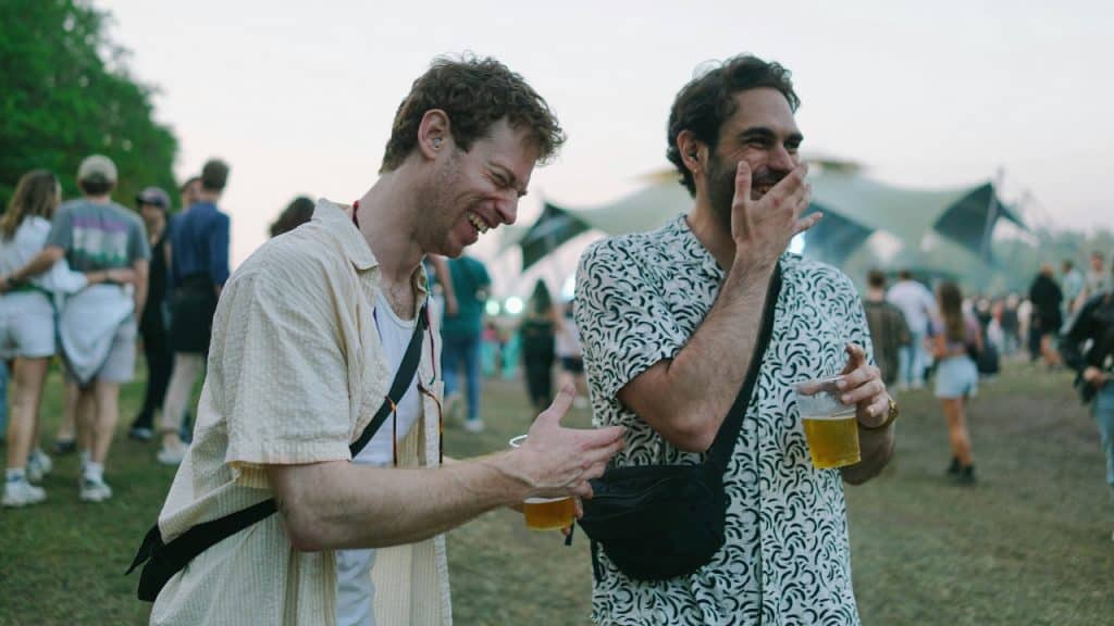 Two men laugh and hold plastic cups while standing in a crowded outdoor field.