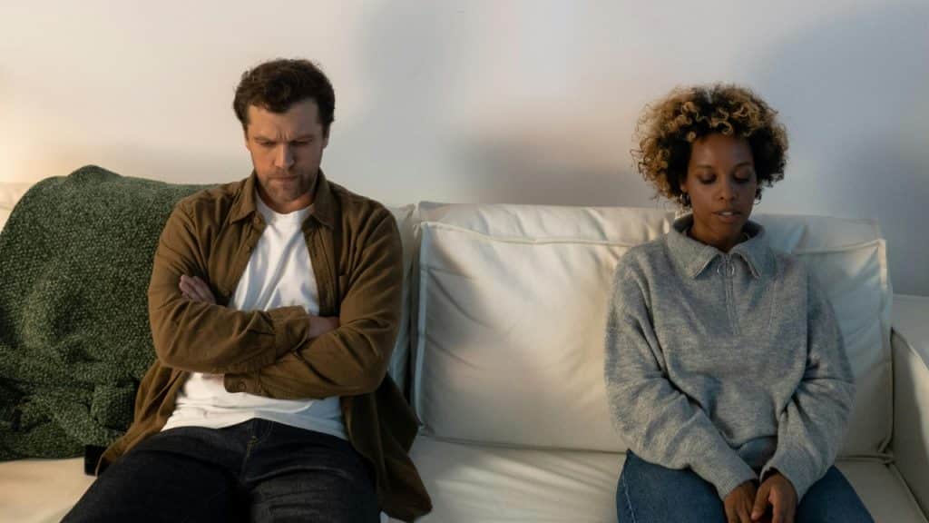 A man and woman sit apart on a white sofa with their eyes looking downward.