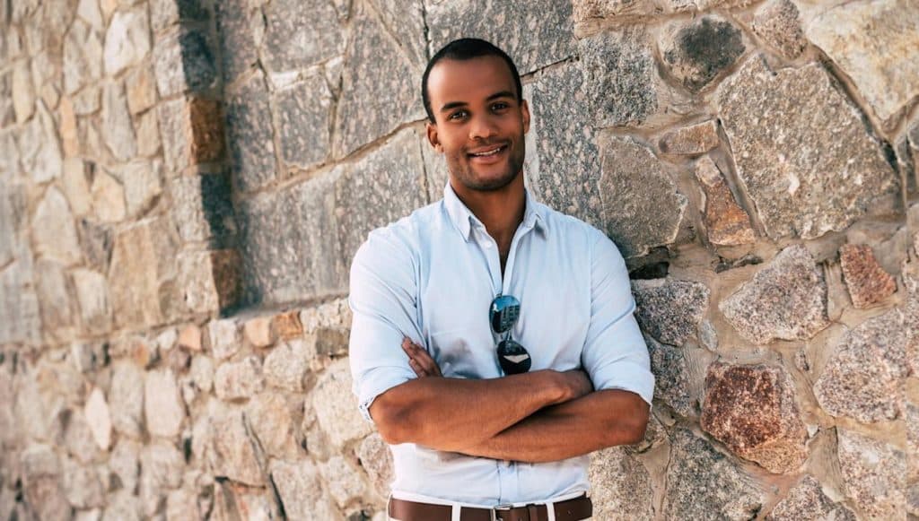 A smiling African man keeping his arms crossed and standing against the stone wall outdoors.