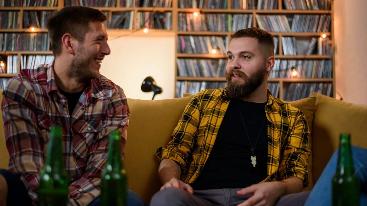 Two men in flannel shirts sit on a couch laughing together in front of bookshelves.