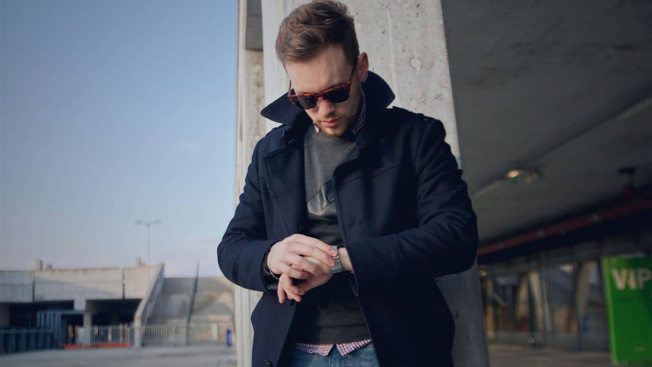 A man in a dark coat and sunglasses checks his watch while leaning against a pillar.