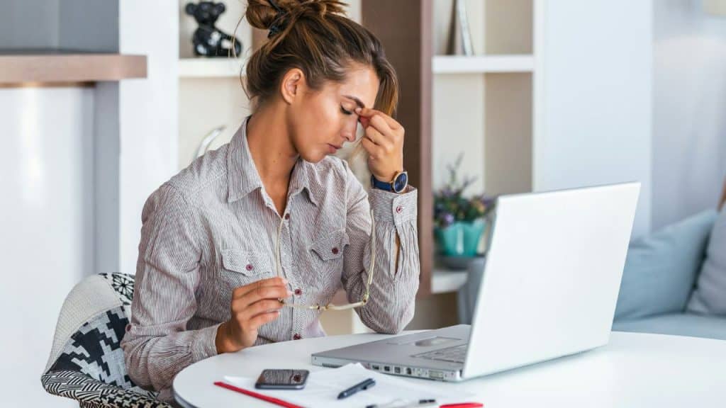 A woman sits at a desk with a laptop, holding glasses and pinching her nose.