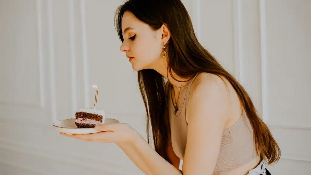 A woman holding a small slice of cake with a lit candle and blowing it out.