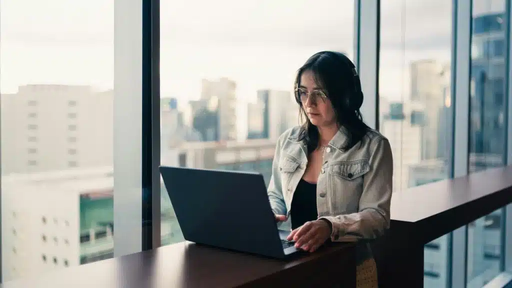 A woman wearing headphones working on a laptop by a window with a city view.