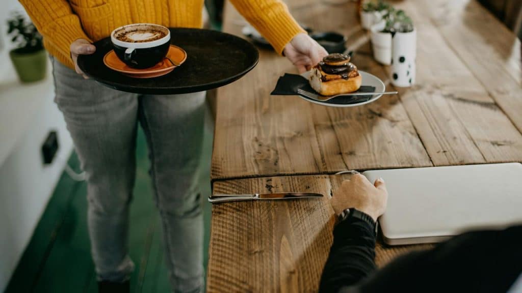 A person serving coffee and a pastry at a wooden table.