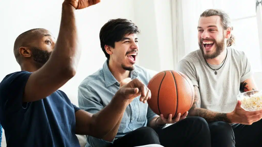 Three men sit together laughing; one holds a basketball and another holds a bowl of popcorn.