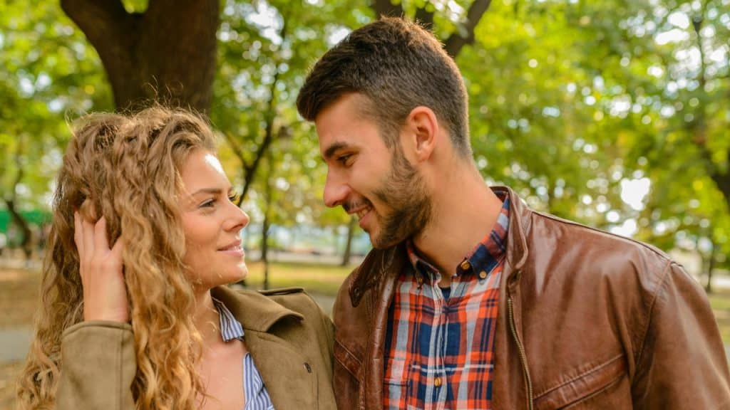 A woman touches her hair while smiling at a man in a park.