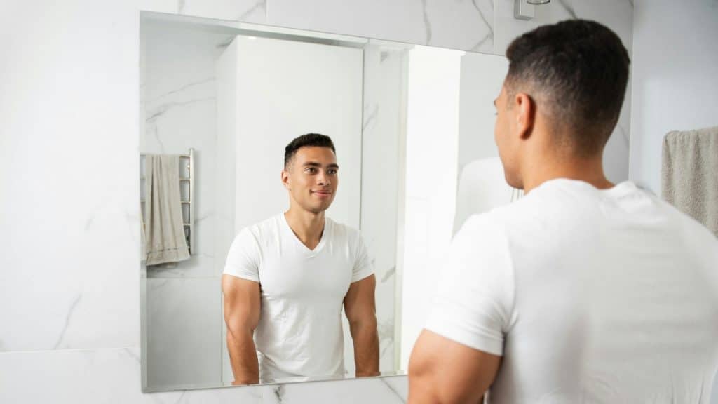 A man in a white v-neck t-shirt looks at his reflection in a bathroom mirror.