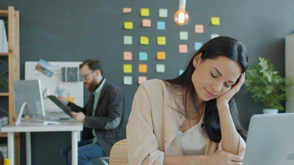 A woman rests her head on her hand at a desk while a man works behind.