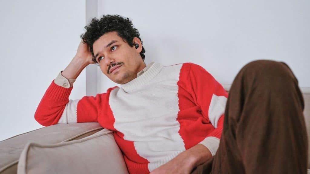 A man with curly hair and a red-and-white sweater reclines on a beige sofa.