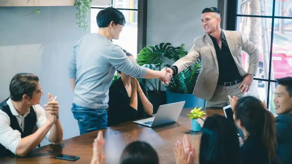 A group of people at a conference table applaud as two men shake hands.