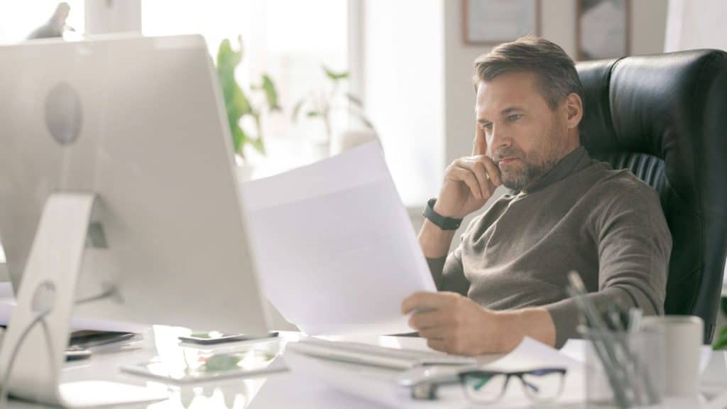 A man sits at a desk and looks at a white paper held in his hand.