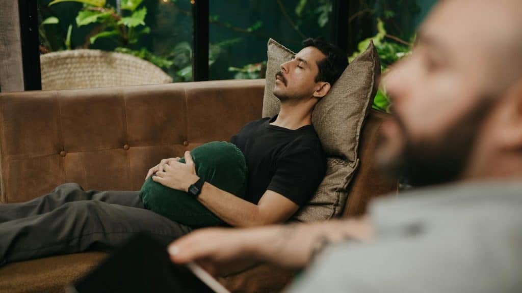 A man resting on a couch with eyes closed, holding a pillow in a calm indoor setting.