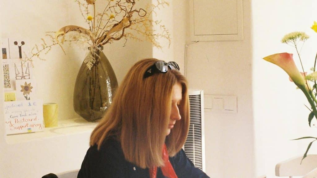 A woman indoors, looking down while seated near decorative vases and flowers.