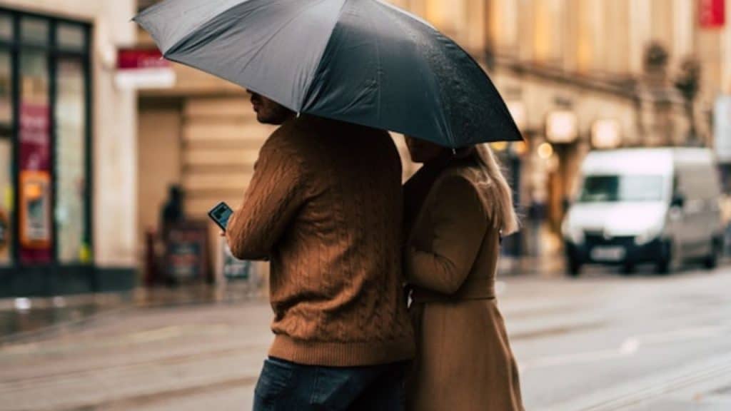 A man and woman walk together on a city street sharing a single large black umbrella.