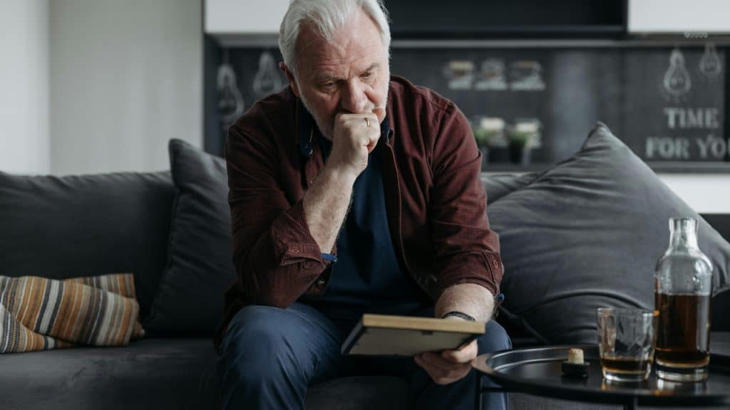 An older man sits on a couch, holding a picture frame and looking distressed.