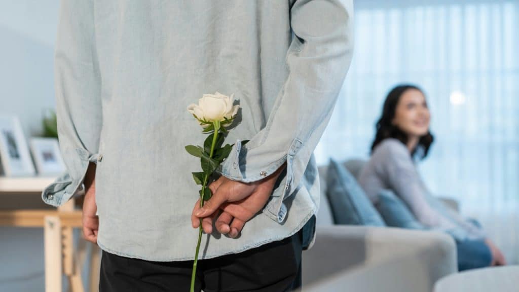 A man hides a single white rose behind his back as a woman sits nearby.