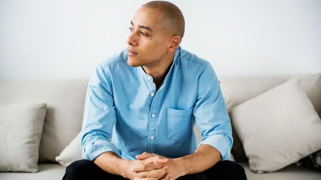 A man in a light blue shirt sits on a couch with his hands clasped, looking sideways.