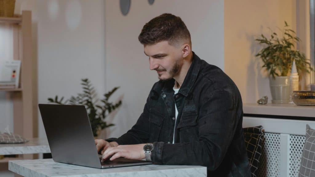 A man types on a laptop while sitting at a table in a room.