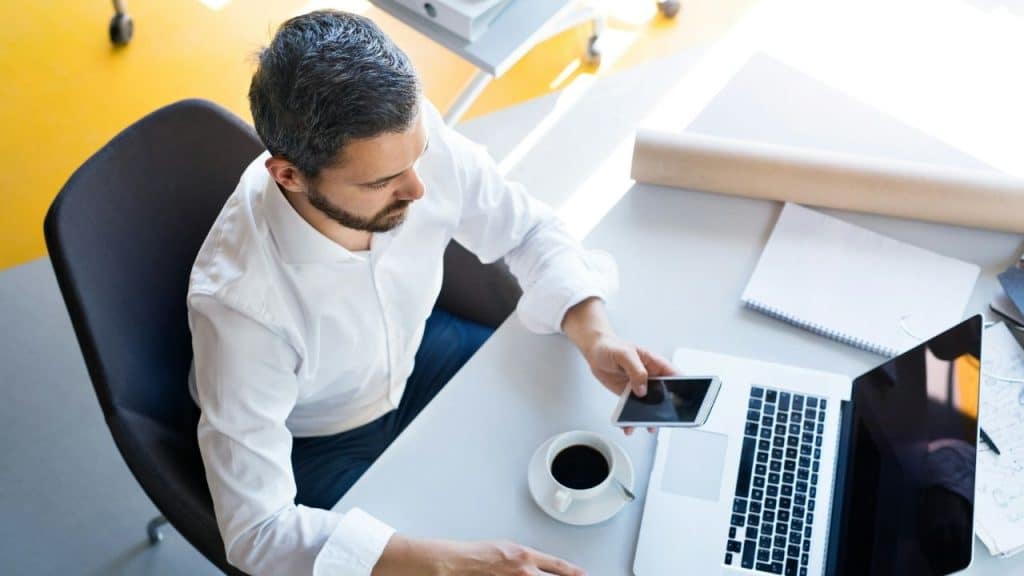 A man in a white shirt sits at a desk using his smartphone near a laptop.