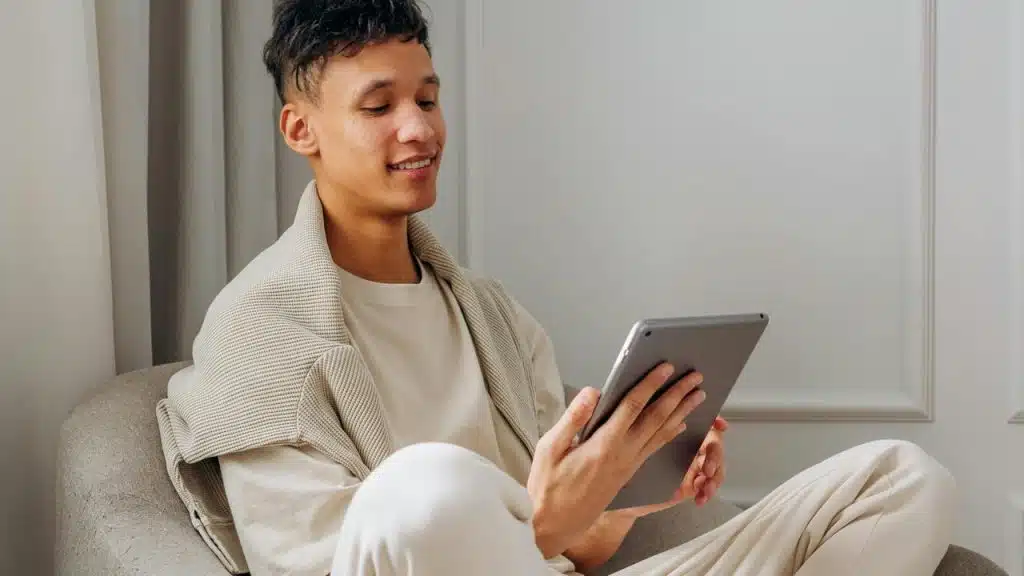 A young man sitting on a couch, smiling while using a tablet indoors.