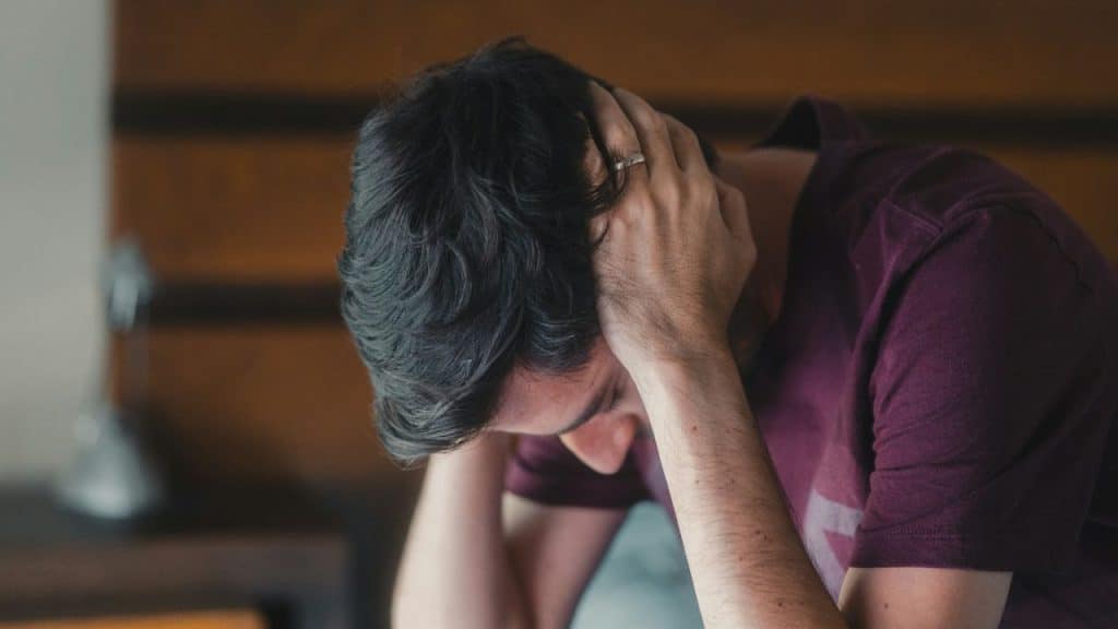 A man with dark hair and a purple shirt rests his head in his hands.