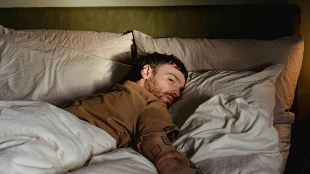 A man with short hair and tattoos lies awake in bed looking to the side.