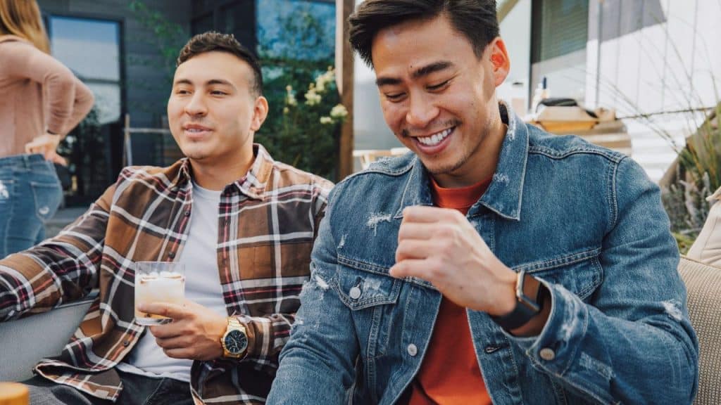 Two men smile while sitting outdoors; one wears a gold watch and the other a smartwatch.