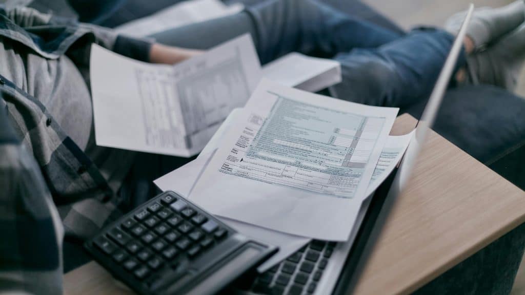 A person sits on a couch reviewing financial tax documents with a calculator nearby.